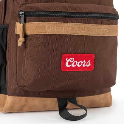 Angled close-up of Coors backpack showing suede zip pull, 'Taste the High Country' suede trim, red Coors patch, and black haul loop over tan suede base
