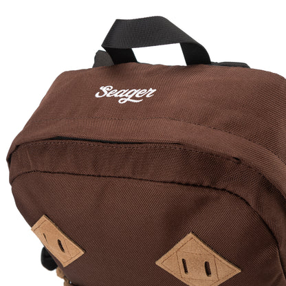 Close-up of the top of a brown backpack showing white embroidered Seager logo, black haul loop, and two tan suede lash tabs.